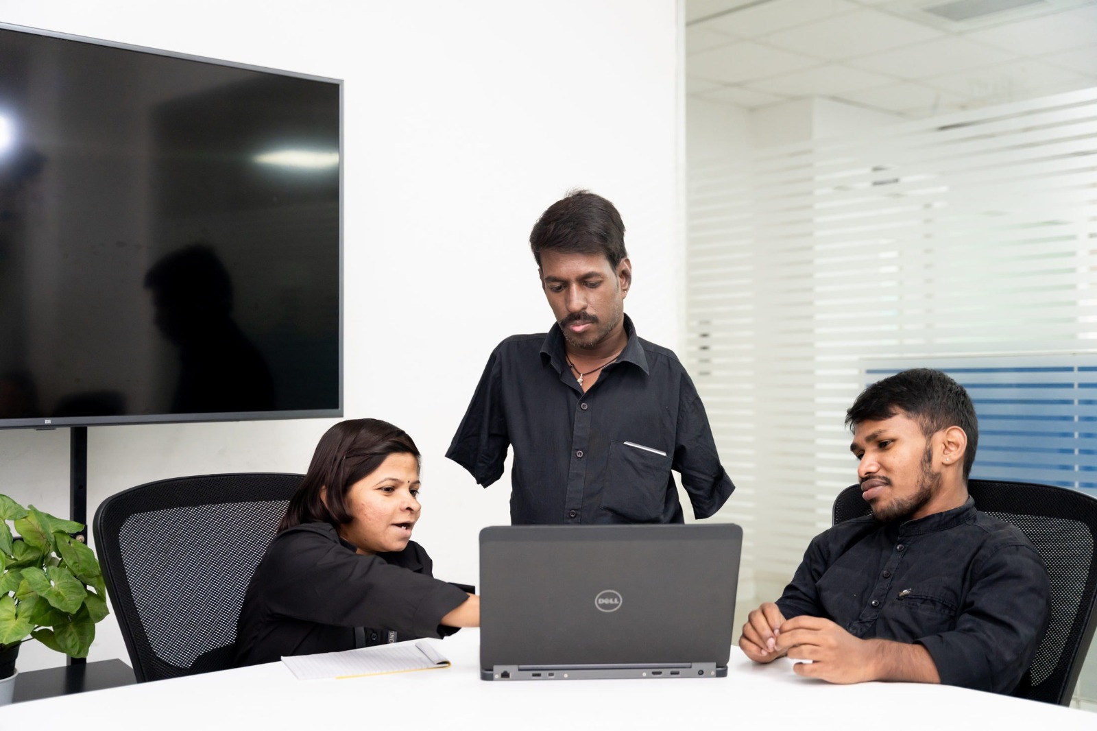 A team of three professionals collaborates around a laptop in a modern office setting. One individual, with a confident and focused expression, gestures towards the screen while explaining a point. Another person attentively observes, standing beside them, while a third team member sits, thoughtfully engaging with the discussion. All are dressed in coordinated black attire, with a potted plant and a large screen in the background adding to the workspace's professional yet welcoming ambiance.