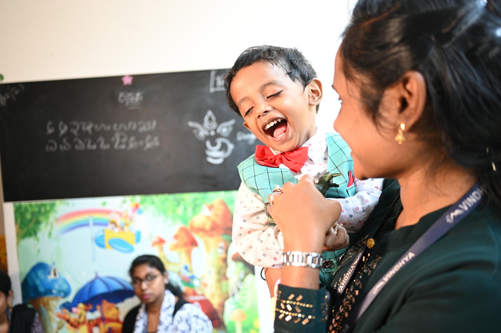 An employee joyfully interacts with a young child at a daycare facility. The child, dressed in a colorful outfit with a red bow tie and green vest, laughs gleefully, creating a heartwarming moment. The background features a vibrant mural with rainbows and whimsical illustrations, alongside a chalkboard with text and drawings. The setting conveys a nurturing and cheerful atmosphere.